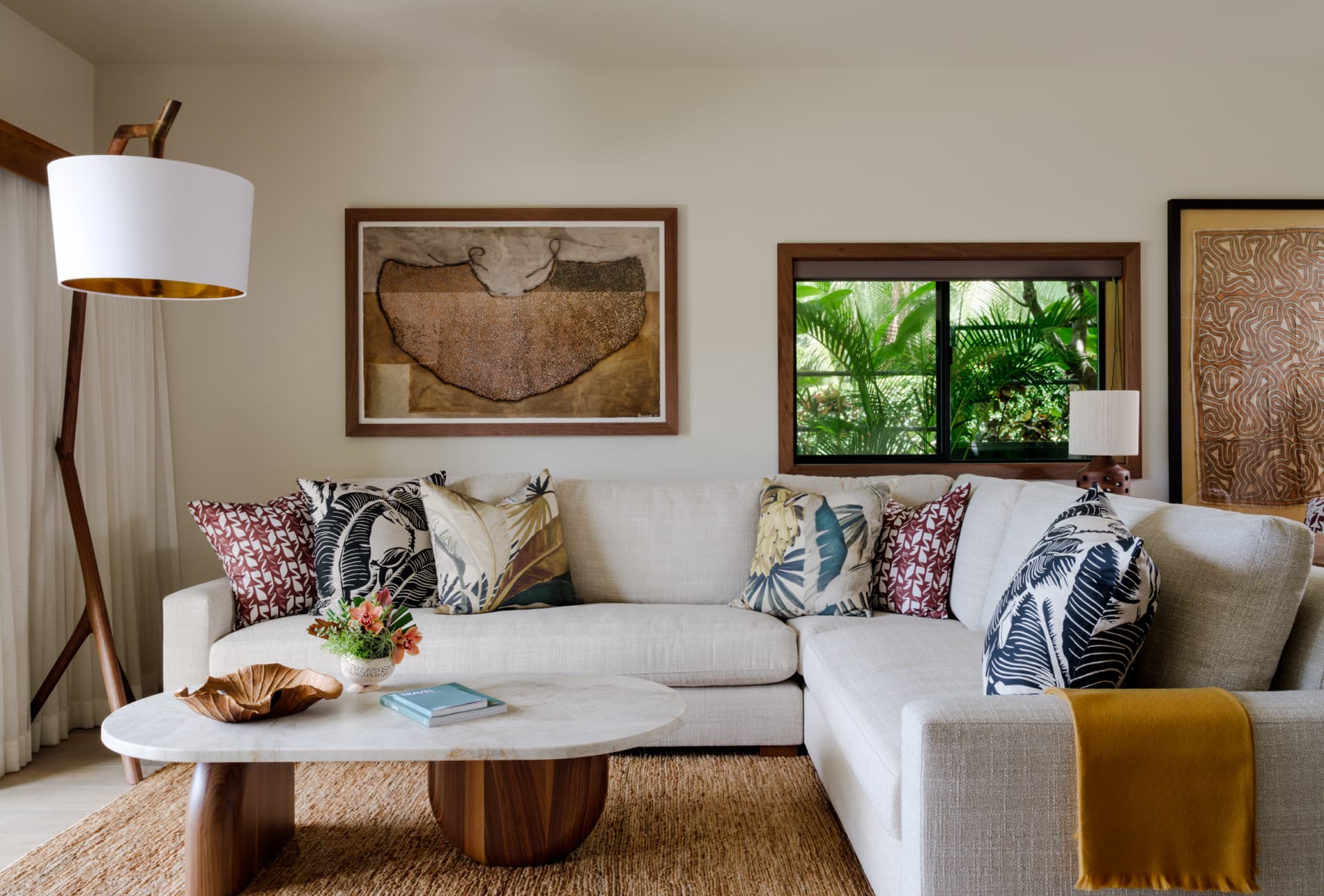 Modern living room with neutral-colored sectional sofa, patterned throw pillows, a wooden coffee table, floor lamp, and wall art. A large window reveals green foliage outside.