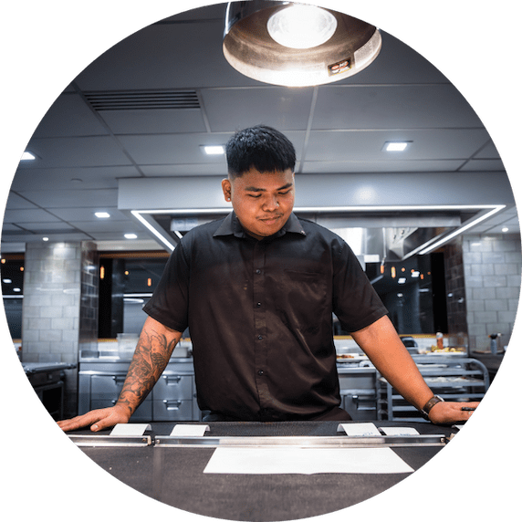 An expo stands focused in a modern kitchen, reviewing the preparation station under bright lighting. Dressed in black, he embodies precision and calm in a refined culinary environment.